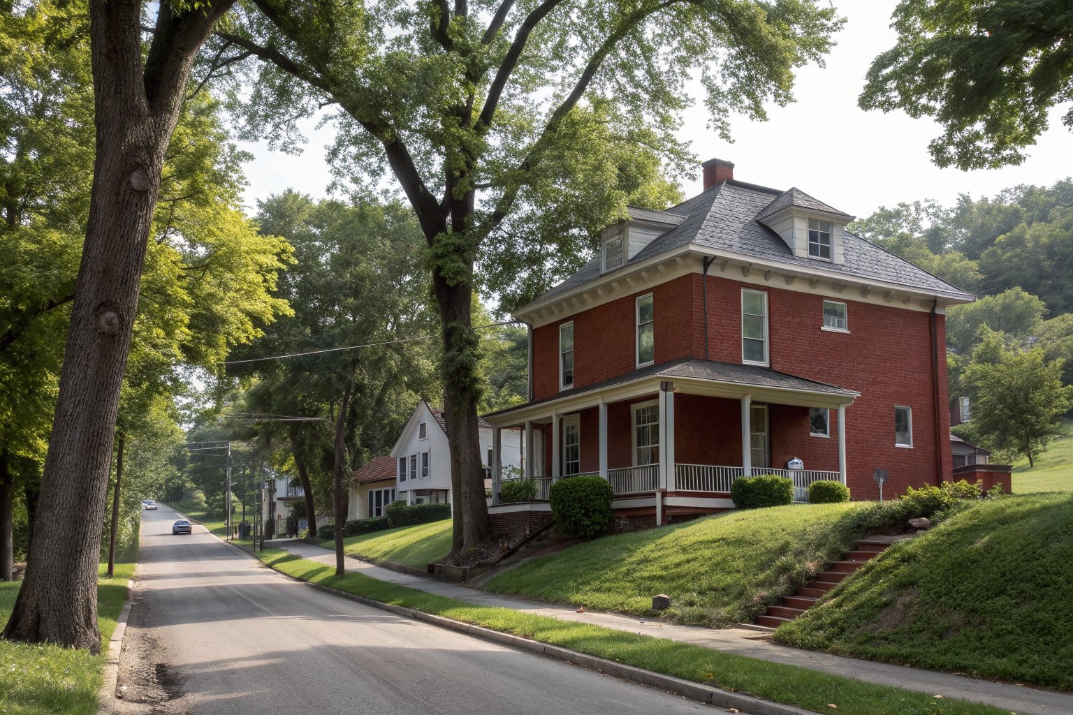 A two-story red brick foursquare house with a gray hipped shingle roof on a sloping street lined with big shade trees