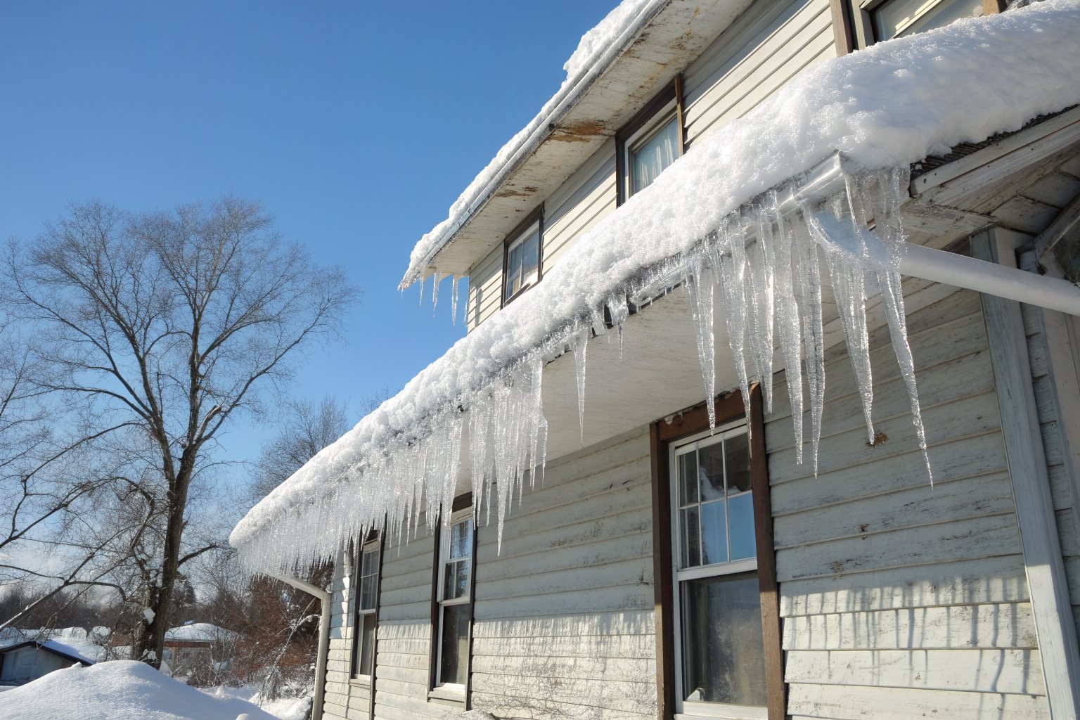 Thick icicles and packed snow hanging from the eaves and gutter of an older two-story house on a sunny winter day