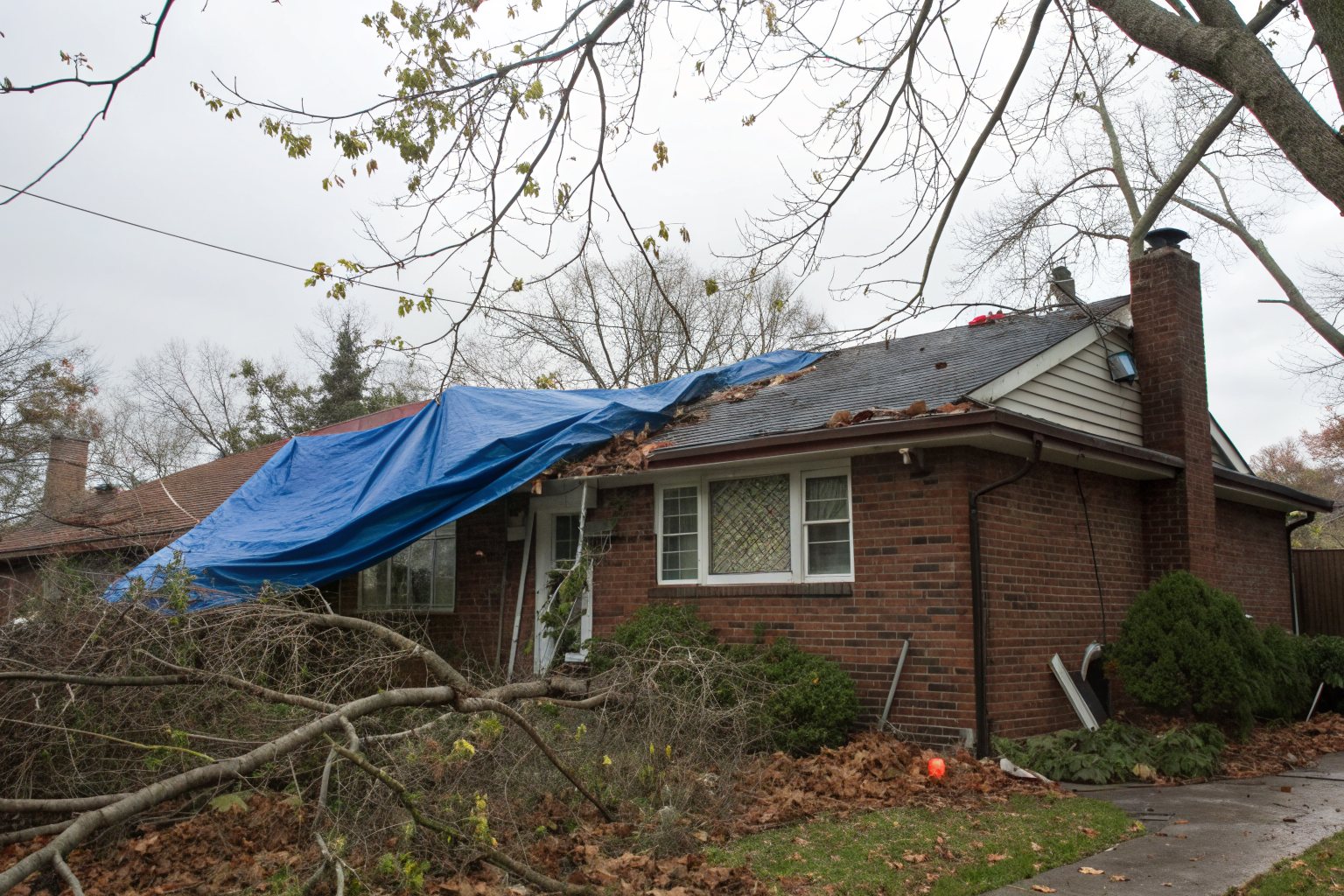 A blue tarp over part of the roof of a brick bungalow, with fallen branches in the yard after a windstorm